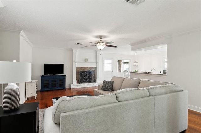 living room featuring ceiling fan, dark wood-type flooring, a brick fireplace, crown molding, and a textured ceiling