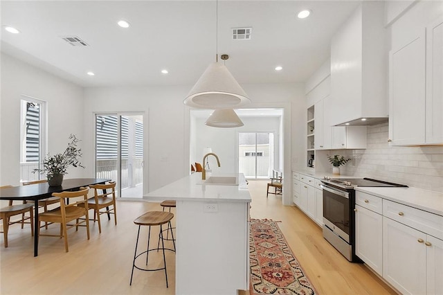 kitchen featuring stainless steel range with electric stovetop, wall chimney exhaust hood, sink, pendant lighting, and an island with sink