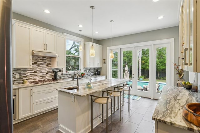 kitchen with white cabinetry, an island with sink, hanging light fixtures, stainless steel gas cooktop, and light stone counters