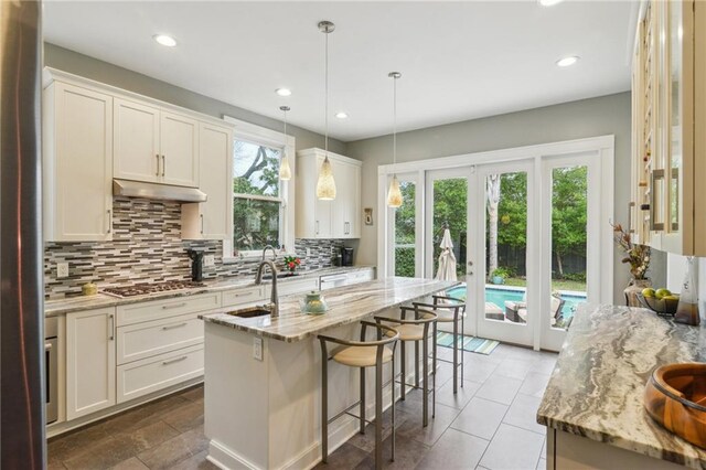 kitchen with light stone countertops, decorative light fixtures, white cabinetry, an island with sink, and stainless steel gas stovetop