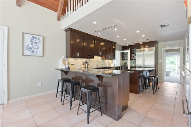 kitchen with a kitchen bar, kitchen peninsula, backsplash, stainless steel built in refrigerator, and dark brown cabinetry