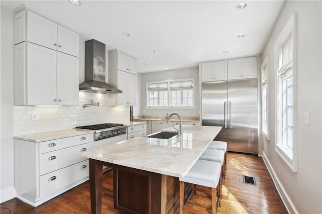 kitchen with wall chimney range hood, a center island with sink, sink, appliances with stainless steel finishes, and white cabinets