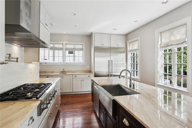 kitchen with light stone countertops, wall chimney exhaust hood, white cabinetry, stainless steel appliances, and sink