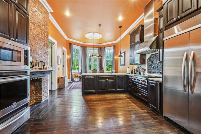 kitchen featuring built in appliances, wall chimney range hood, dark brown cabinets, and kitchen peninsula