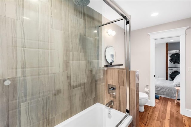 bathroom with hardwood / wood-style flooring, stacked washing maching and dryer, shower / bath combination with glass door, and toilet