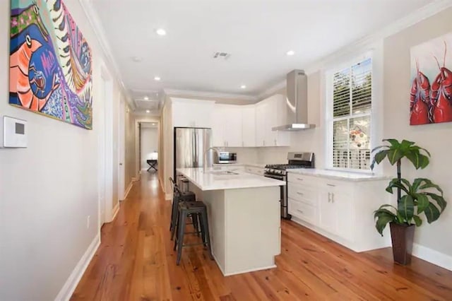 kitchen with stainless steel appliances, white cabinets, wall chimney exhaust hood, a breakfast bar area, and a kitchen island with sink