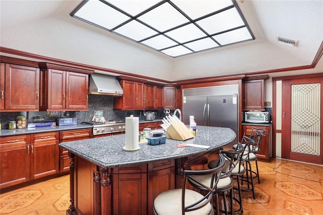 kitchen with wall chimney exhaust hood, a kitchen island, backsplash, dark stone counters, and appliances with stainless steel finishes