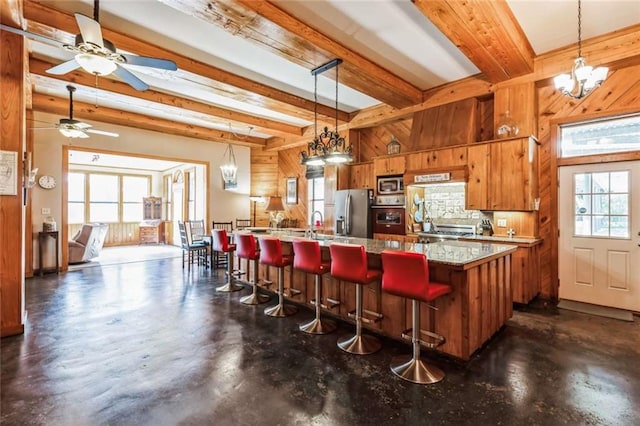 kitchen with wood walls, stainless steel appliances, beam ceiling, hanging light fixtures, and ceiling fan with notable chandelier