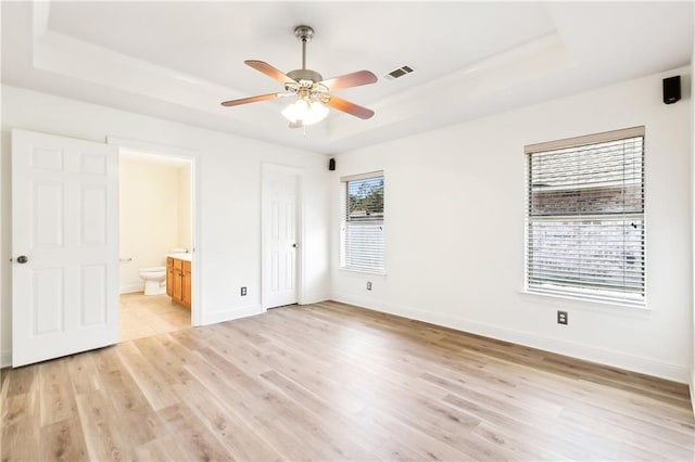 unfurnished bedroom featuring a raised ceiling, ensuite bath, ceiling fan, and light wood-type flooring