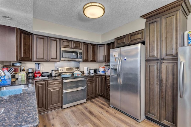 kitchen with appliances with stainless steel finishes, light wood-type flooring, dark stone countertops, a textured ceiling, and dark brown cabinetry