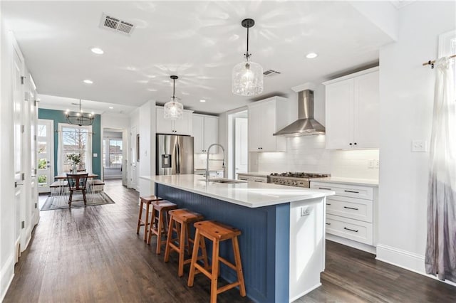 kitchen with an island with sink, appliances with stainless steel finishes, wall chimney exhaust hood, white cabinets, and sink