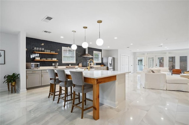 kitchen with pendant lighting, appliances with stainless steel finishes, wall chimney exhaust hood, an island with sink, and gray cabinets