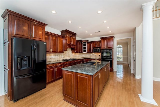 kitchen with dark stone countertops, a center island with sink, black appliances, sink, and ornate columns