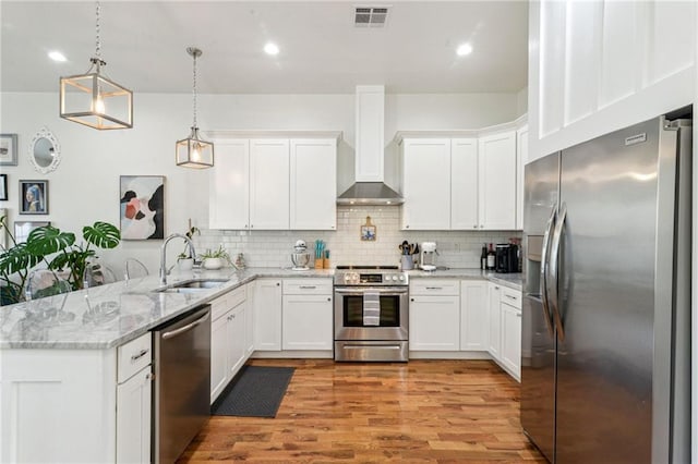 kitchen featuring appliances with stainless steel finishes, white cabinets, wall chimney exhaust hood, hanging light fixtures, and kitchen peninsula