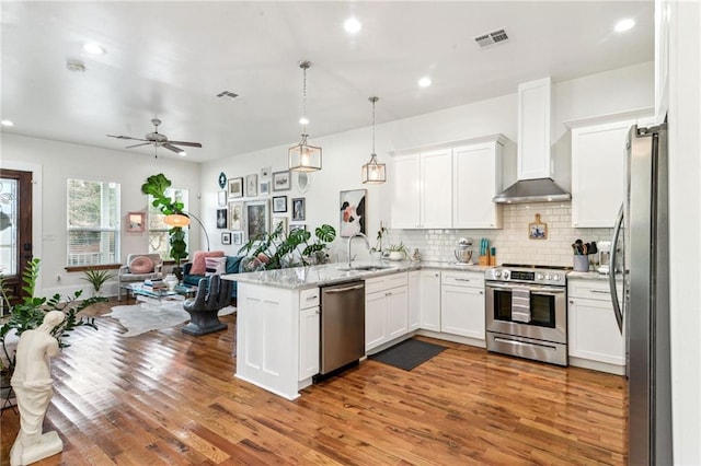 kitchen with decorative light fixtures, kitchen peninsula, sink, appliances with stainless steel finishes, and wall chimney exhaust hood