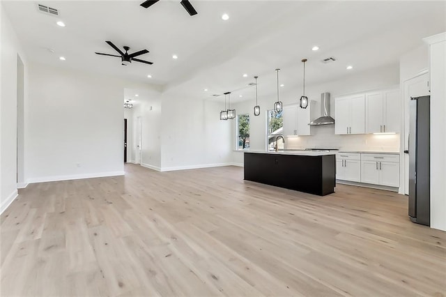 kitchen with white cabinetry, wall chimney range hood, hanging light fixtures, stainless steel fridge, and a kitchen island with sink