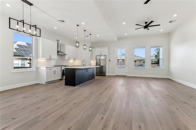 kitchen featuring an island with sink, stainless steel appliances, hanging light fixtures, wall chimney range hood, and white cabinets