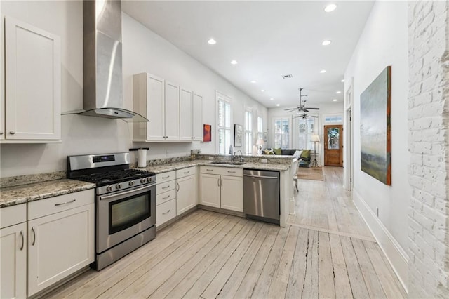 kitchen with sink, light stone counters, appliances with stainless steel finishes, kitchen peninsula, and wall chimney range hood