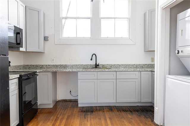 kitchen featuring black appliances, a sink, light stone counters, dark wood finished floors, and stacked washer / drying machine