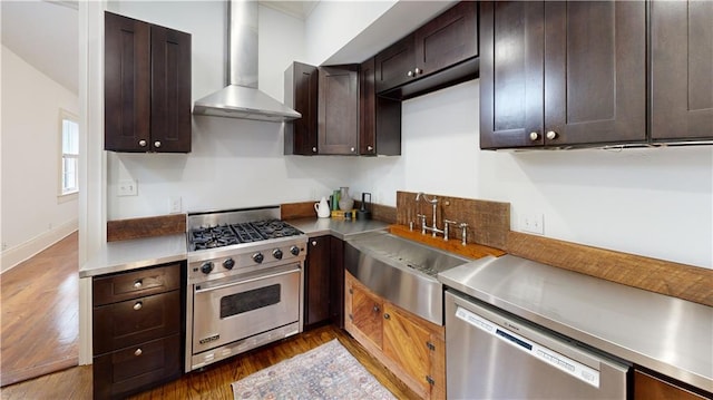 kitchen featuring appliances with stainless steel finishes, stainless steel counters, dark brown cabinets, dark wood-type flooring, and wall chimney range hood