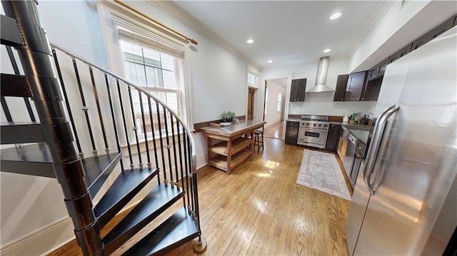 kitchen with appliances with stainless steel finishes, dark brown cabinetry, ornamental molding, light hardwood / wood-style floors, and wall chimney exhaust hood