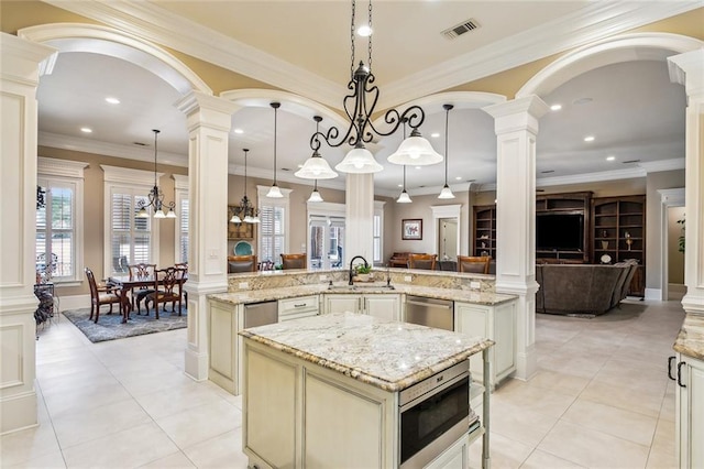 kitchen featuring a kitchen island with sink, hanging light fixtures, cream cabinets, and decorative columns