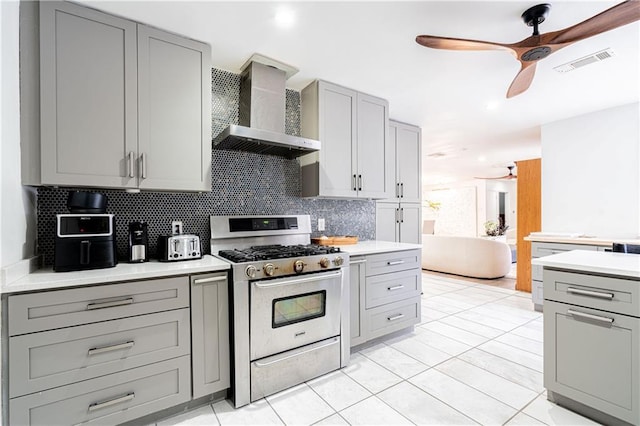 kitchen with gray cabinetry, stainless steel gas stove, ceiling fan, wall chimney range hood, and backsplash