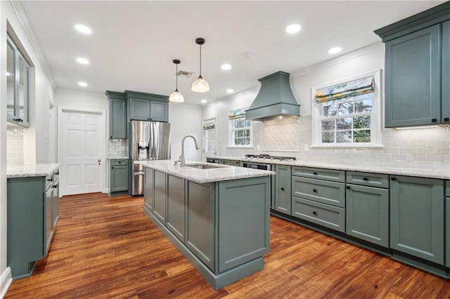 kitchen featuring sink, premium range hood, stainless steel appliances, light stone countertops, and an island with sink