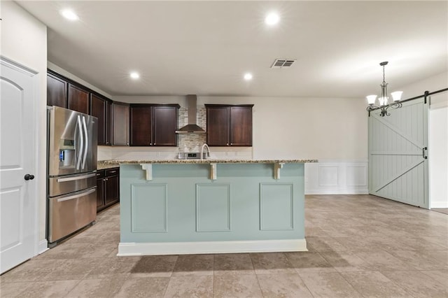 kitchen with light stone counters, wall chimney exhaust hood, a barn door, and stainless steel fridge with ice dispenser