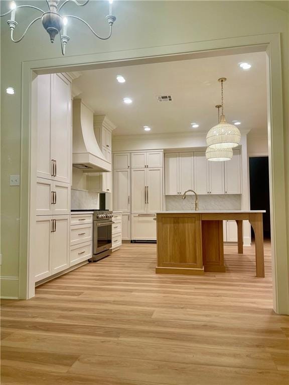 kitchen with stainless steel stove, hanging light fixtures, custom range hood, an island with sink, and white cabinets