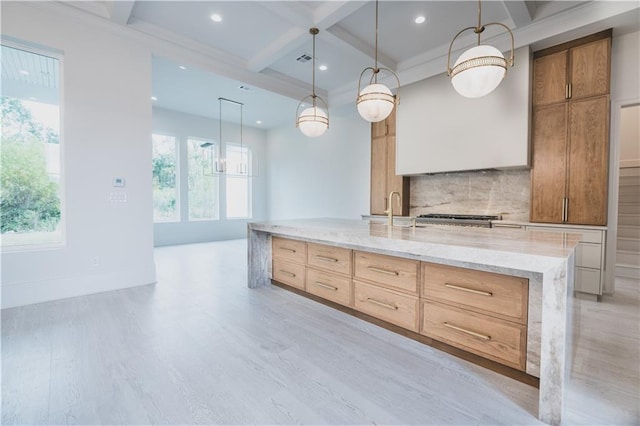 kitchen featuring decorative light fixtures, decorative backsplash, coffered ceiling, a kitchen island with sink, and light stone countertops