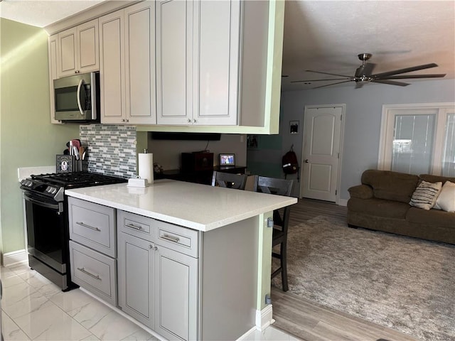 kitchen featuring a breakfast bar area, gray cabinets, ceiling fan, gas range oven, and backsplash