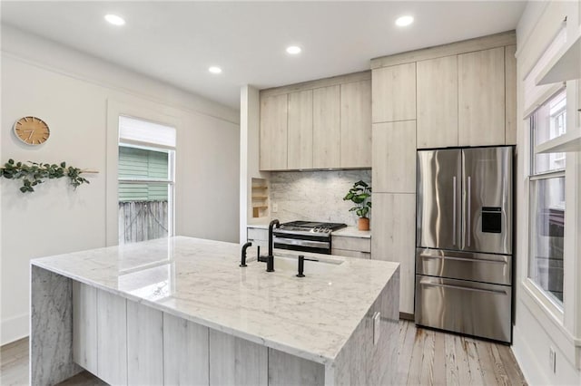 kitchen featuring appliances with stainless steel finishes, backsplash, light stone countertops, an island with sink, and light wood-type flooring