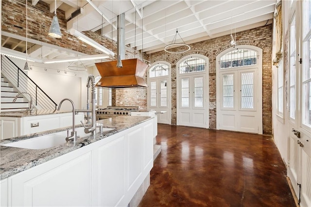 kitchen featuring brick wall, sink, white cabinets, light stone counters, and custom range hood