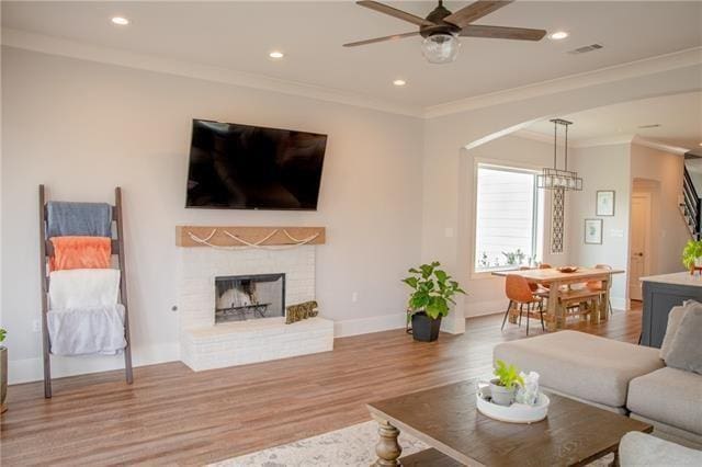 living room with ornamental molding, wood-type flooring, ceiling fan with notable chandelier, and a brick fireplace