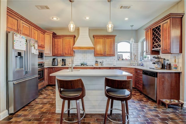 kitchen featuring pendant lighting, appliances with stainless steel finishes, tasteful backsplash, custom range hood, and a center island with sink