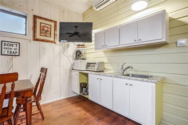 kitchen with white microwave, white cabinetry, wood walls, a sink, and wood finished floors