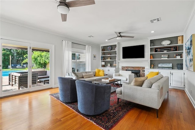 living room with a brick fireplace, light hardwood / wood-style flooring, ornamental molding, built in features, and ceiling fan