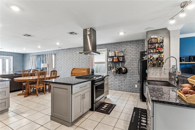 kitchen with light tile patterned floors, gray cabinets, stainless steel gas stove, island range hood, and brick wall