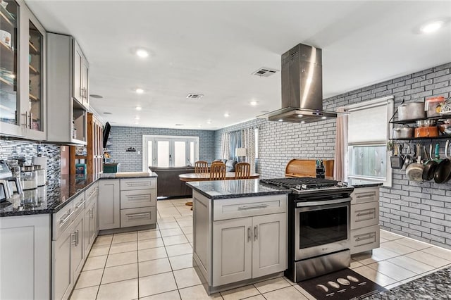 kitchen with gray cabinets, light tile patterned flooring, stainless steel gas stove, dark stone countertops, and island exhaust hood