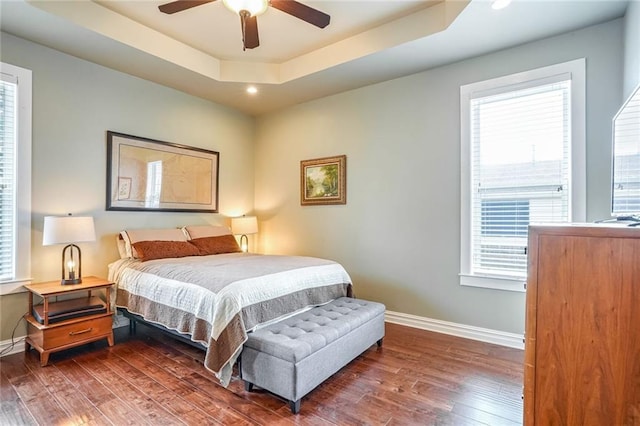 bedroom featuring dark hardwood / wood-style floors, ceiling fan, a tray ceiling, and multiple windows