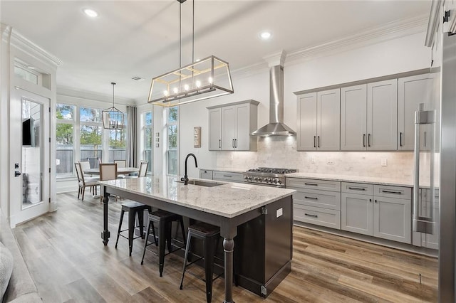 kitchen featuring pendant lighting, sink, wall chimney range hood, light stone counters, and a center island with sink