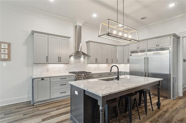 kitchen with wall chimney exhaust hood, sink, high end appliances, light stone counters, and an island with sink