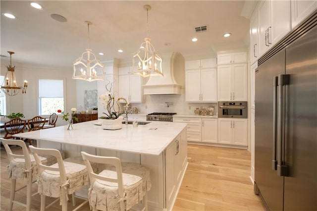 kitchen with hanging light fixtures, stainless steel appliances, an island with sink, and custom range hood