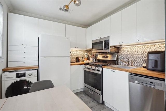 kitchen with appliances with stainless steel finishes, butcher block counters, tasteful backsplash, white cabinets, and washer / dryer