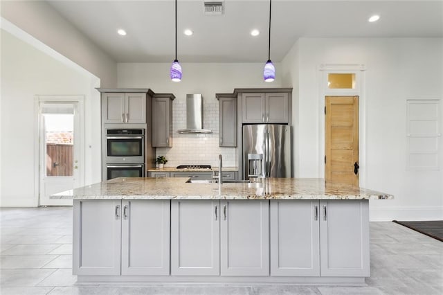 kitchen with appliances with stainless steel finishes, wall chimney exhaust hood, light stone counters, decorative light fixtures, and a large island with sink