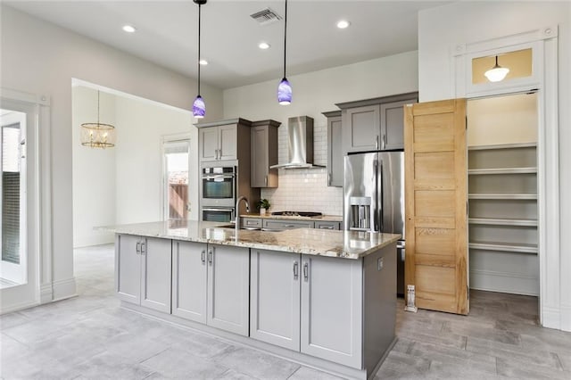 kitchen with wall chimney exhaust hood, light stone counters, appliances with stainless steel finishes, pendant lighting, and a kitchen island with sink