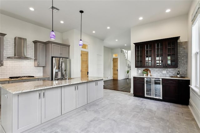 kitchen with wine cooler, decorative light fixtures, appliances with stainless steel finishes, an island with sink, and wall chimney range hood