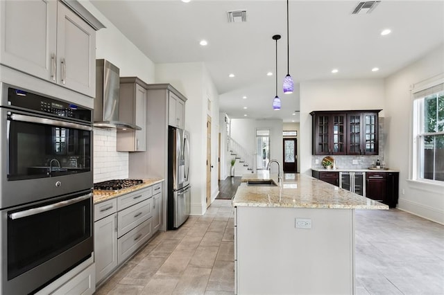 kitchen featuring wall chimney exhaust hood, sink, a center island with sink, gray cabinets, and stainless steel appliances