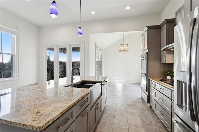 kitchen with sink, an island with sink, pendant lighting, stainless steel appliances, and light stone countertops